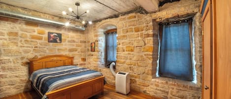 A bedroom with a wooden bed, stone walls, a window with curtains, and a ceiling fan.