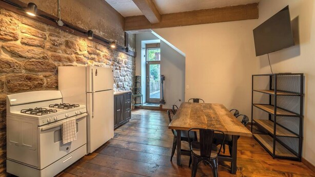 A kitchen with a stone wall, a stove, a refrigerator, and a wooden dining table with chairs.