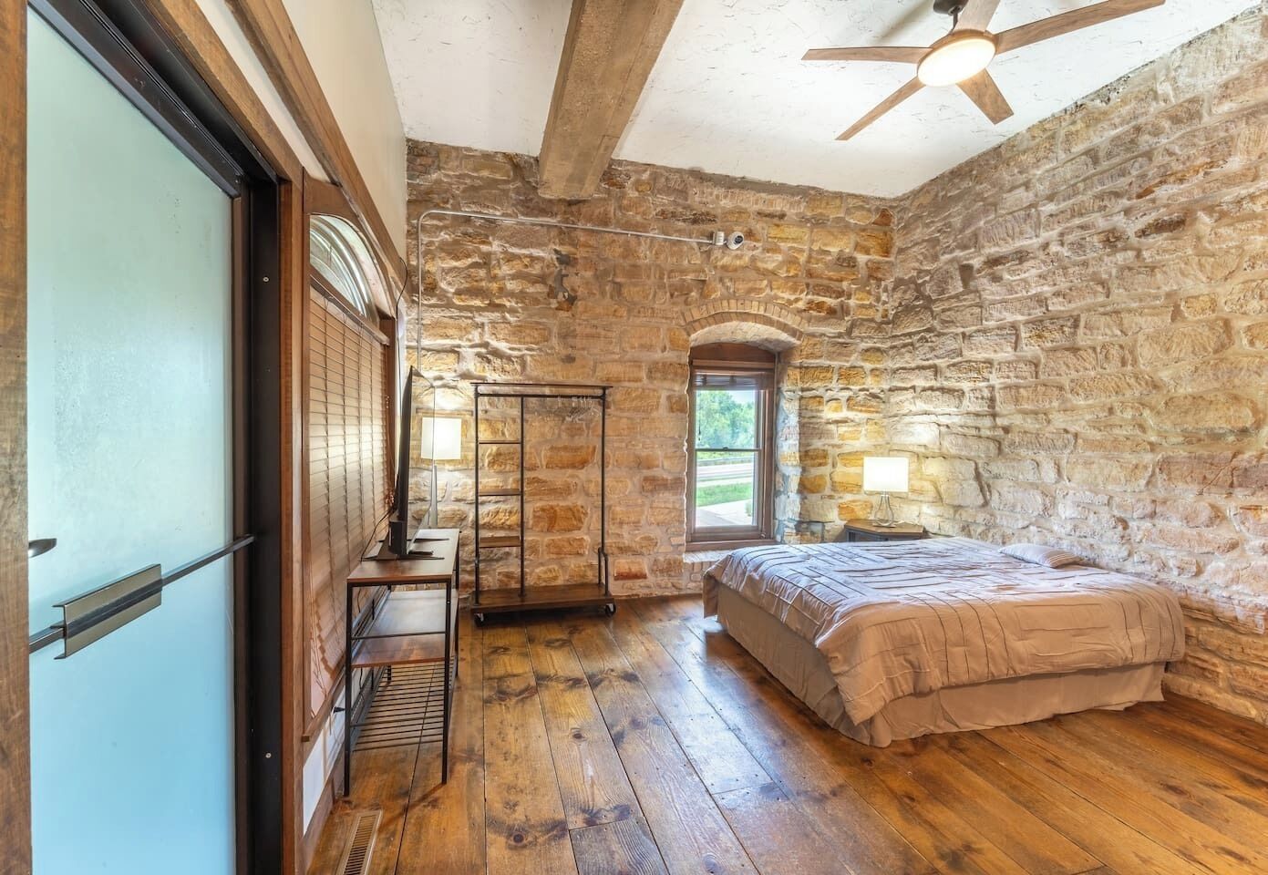 Een slaapkamer met stenen muren, een houten vloer, een bed, een bureau en een raam.