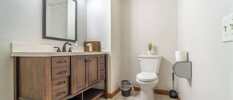 A bathroom with a vanity, mirror, toilet, and a trash bin.