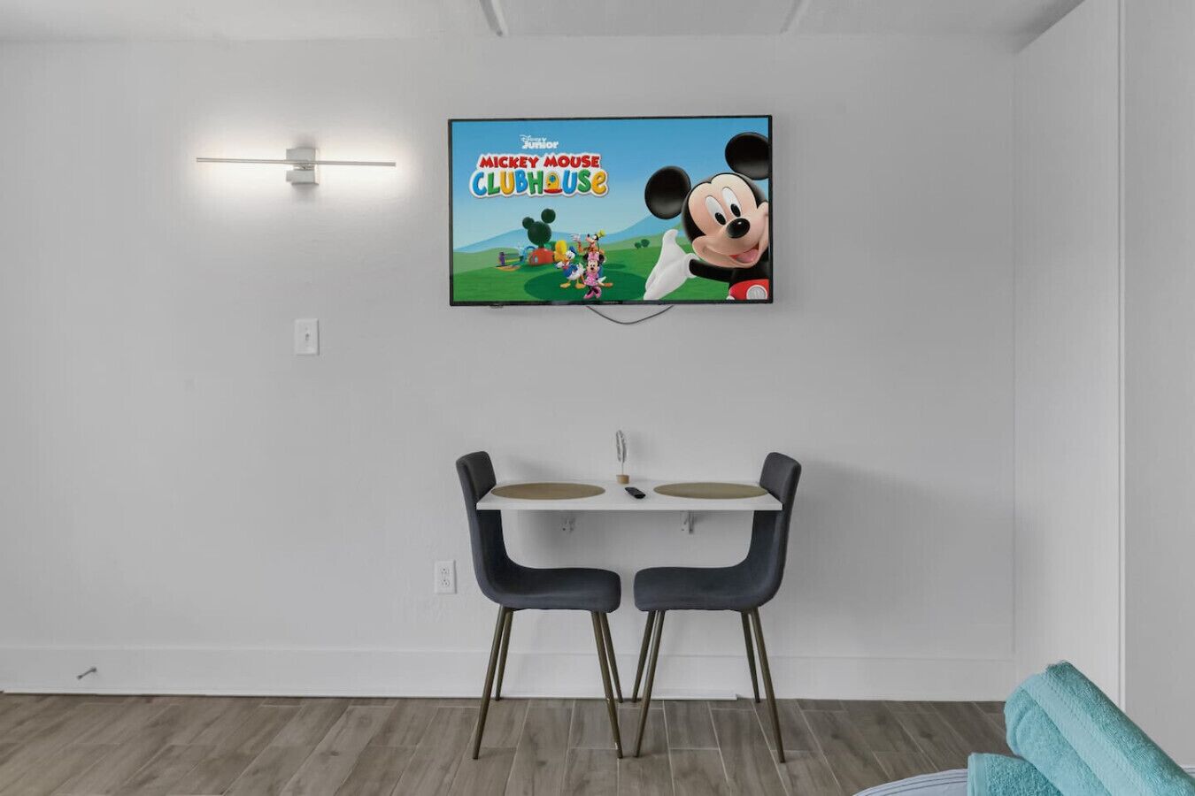 En moderne stue med et fladskærms-tv på væggen, der viser 'Mickey Mouse Klubben'. Der er et spisebord med to stole foran.