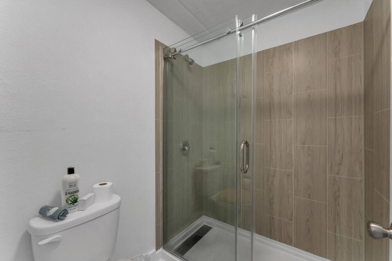 A glass-enclosed shower with a wooden panel wall, a toilet with a toilet paper holder, and a bottle of shampoo.