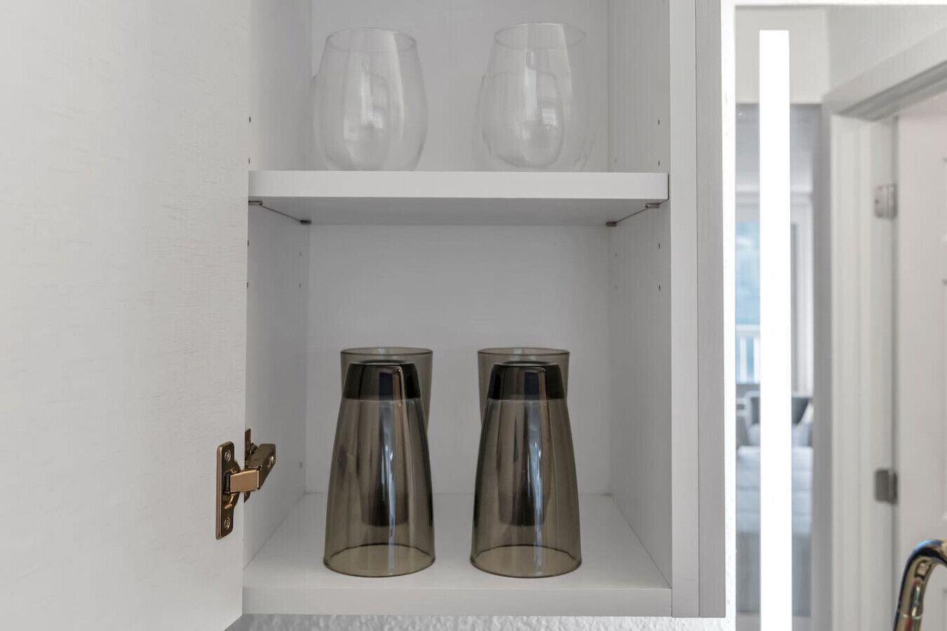 A white cabinet with two shelves; the top shelf holds two clear glass cups, and the bottom shelf has two clear glass pitchers.