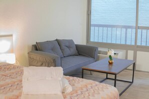 Ein Zimmer mit einem Bett, einem grauen Sofa, einem kleinen Tisch mit einer Vase und einem Fenster mit Meerblick.