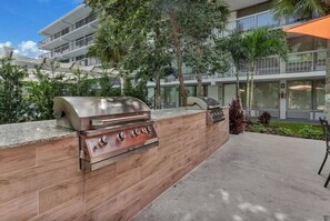 Ein Außenbereich mit Küche, der einen eingebauten Grill und Backofen sowie eine Betonterrasse und viel Grün umfasst.