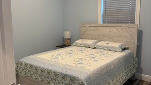 Une chambre à coucher comprenant un lit, un ventilateur de plafond, une fenêtre avec des stores, une table de chevet avec une lampe et une porte.