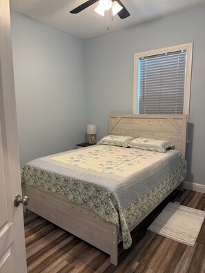 A bedroom with a bed, a ceiling fan, a window with blinds, a nightstand with a lamp, and a door. - Furnished 3 bedroom, easy I-4 Access (Plant City)