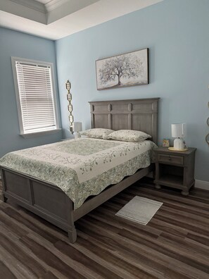 A bedroom with a bed, a nightstand, a wall-mounted lamp, a window with blinds, and a painting of a tree. - Furnished 3 bedroom, easy I-4 Access (Plant City)