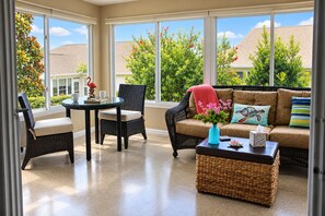 A sunroom with a glass door, a round table, two wicker chairs, a sofa with cushions, and a small table with a vase and a tissue box.