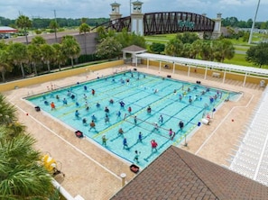 A swimming pool with multiple lanes, surrounded by a covered area with a roof, and a bridge in the background.