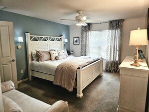 Une chambre à coucher avec un lit, un ventilateur de plafond, une table de chevet avec une lampe et une fenêtre avec des rideaux.