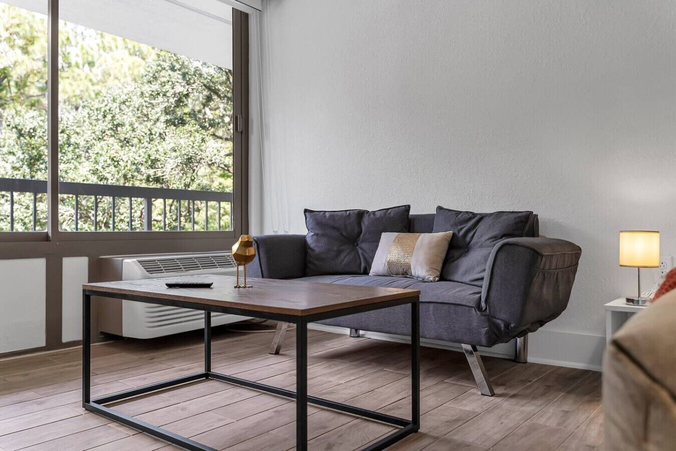 En moderne stue med en grå sofa, et træbord og et vindue med udsigt til grønne områder.