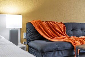 A hotel room with a bed, a bedside table, a lamp, and a sofa with an orange blanket. - Chic Disney Getaway + Free Parking | Orl (Kissimmee)