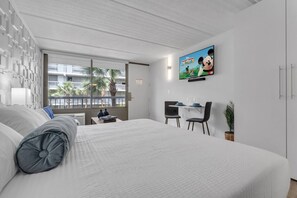 A modern hotel room with a large bed, a desk with two chairs, a television mounted on the wall, and a window with a view of palm trees. - Modern Oasis Near Disney + Free Parking | Orl (Kissimmee)
