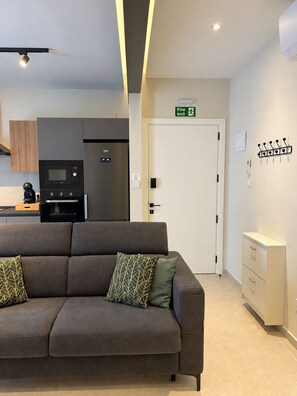 A modern living room with a sofa, a coffee machine, a microwave, and a refrigerator. There is a door with a fire exit sign and a coat rack on the wall.
