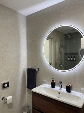 A modern bathroom with a round mirror, a white sink, a dark wooden vanity, a towel rack, and a glass shower enclosure.