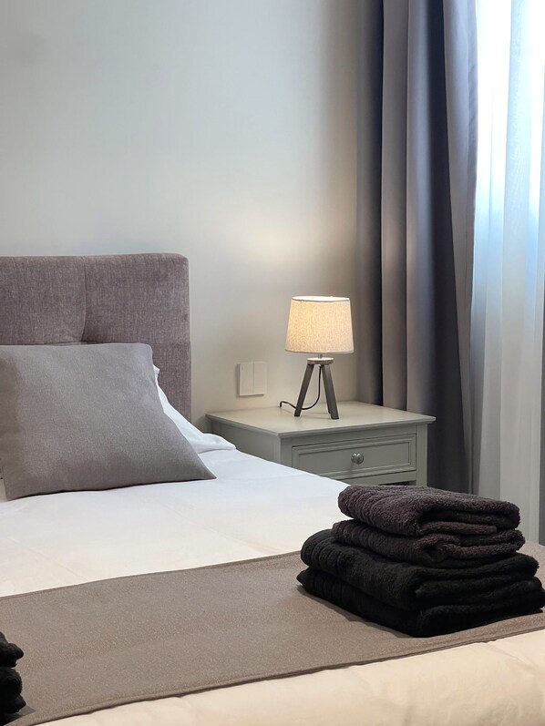 A neatly made bed with a grey headboard, a bedside lamp, and a stack of folded towels.