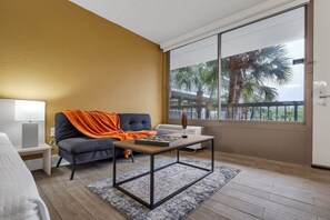 A modern hotel room with a sofa, a small table, a lamp, and a large window with a view of palm trees. - Elegant Disney Escape + Pool & Parking | Orl (Kissimmee)