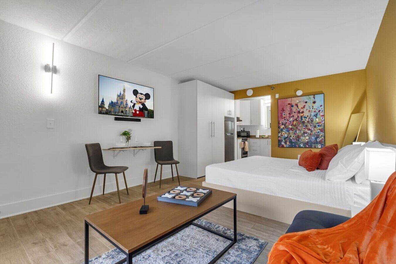 A modern hotel room with a bed, a desk, two chairs, a coffee table, and a painting on the wall.