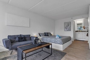 Ein modernes, kompaktes Wohnzimmer mit Sofa, Bett mit gemusterten Bettwaren, kleinem Tisch und einer Küchenzeile.