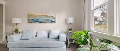 Un salon comprenant un canapé, deux tables d’appoint avec des lampes, un tableau accroché au mur et une grande fenêtre avec des rideaux.