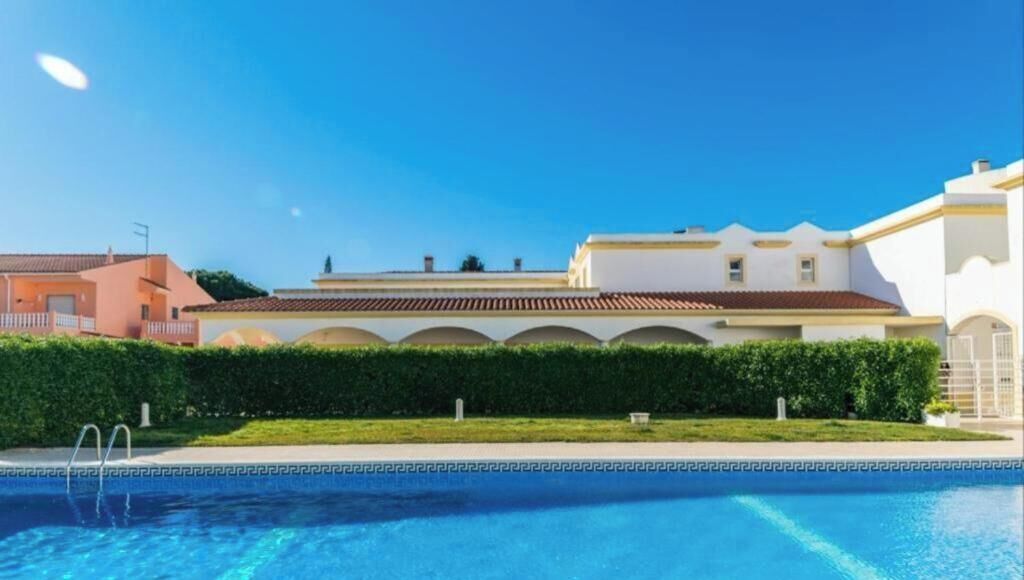 Une piscine, avec un ciel bleu dégagé au-dessus, entourée d’un beau jardin bien entretenu et d’un bâtiment doté d’un toit en tuiles.