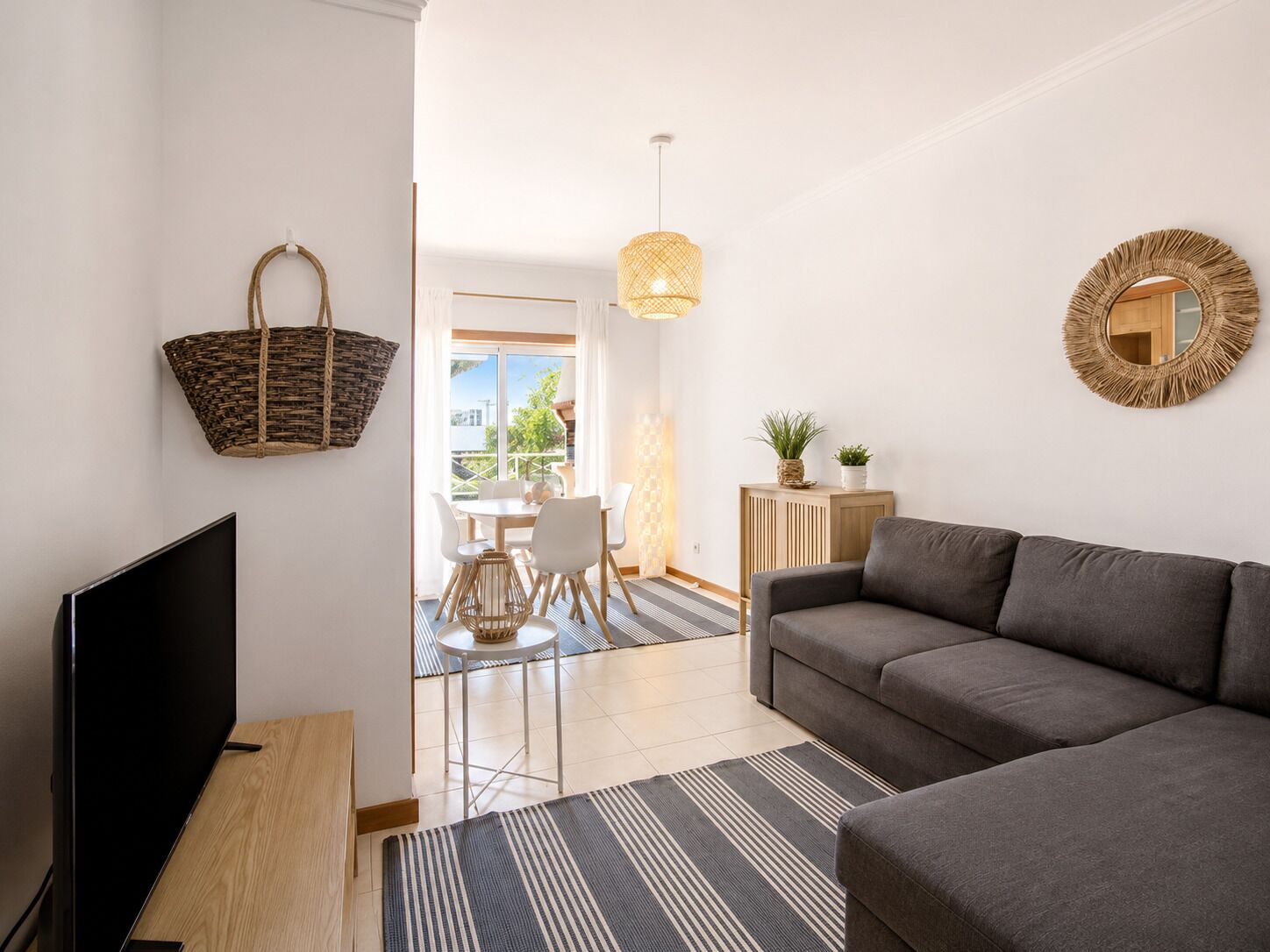 A modern living room with a flat-screen TV, a grey sofa, a woven basket, a round mirror, and a dining area with natural light.