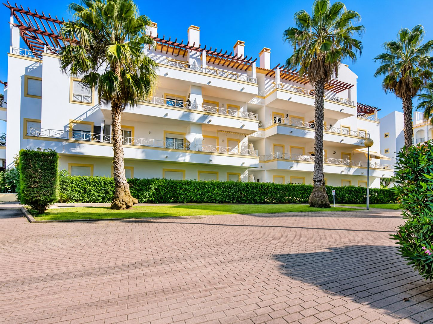 Un hotel su più piani con balconi, palme e un vialetto lastricato.