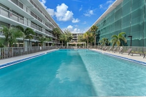 Una piscina de hotel con agua azul cristalina, rodeada por una cerca blanca y un edificio con balcones en un lateral.