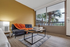 A modern hotel room with a sofa, a small table, a lamp, and a large window with a view of palm trees. - Disney Getaway | Cozy Studio, Pool & Free Parking (Kissimmee)