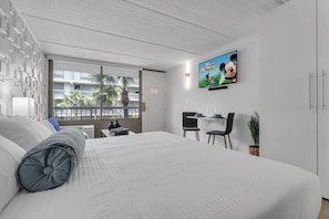 A hotel room with a large bed, a TV mounted on the wall, a small table with chairs, and a window with a view of palm trees. - Studio Getaway Near Disney + Free Parking (Kissimmee)