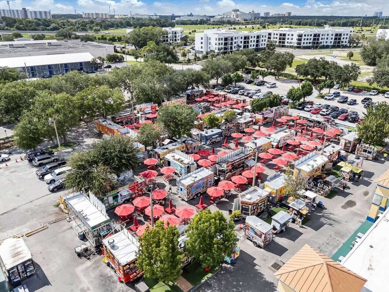 An aerial view of a parking lot with multiple food trucks and red umbrellas, surrounded by trees and buildings.