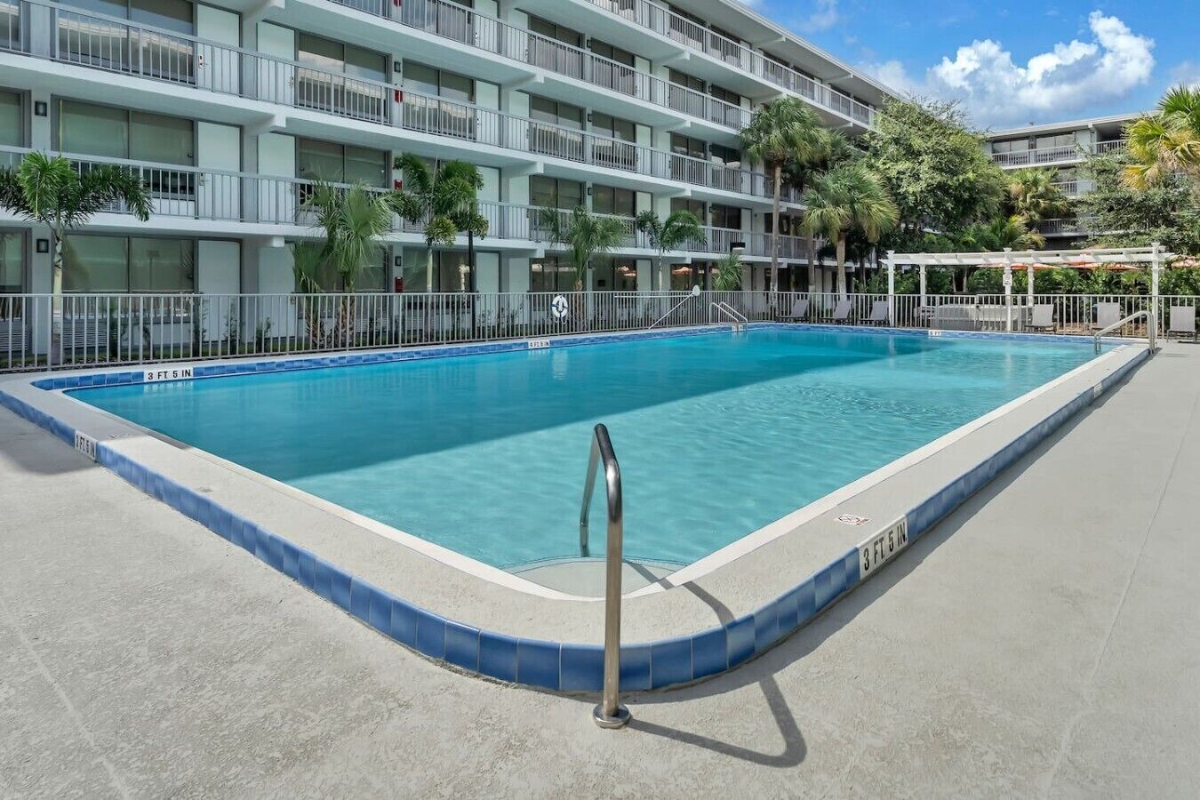 Una piscina rectangular con borde azul, rodeada por una solana de hormigón, y un edificio de varios pisos con balcones al fondo.