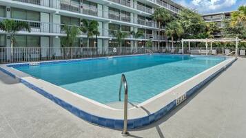 Una piscina rectangular con borde azul, rodeada por una solana de hormigón, y un edificio de varios pisos con balcones al fondo.