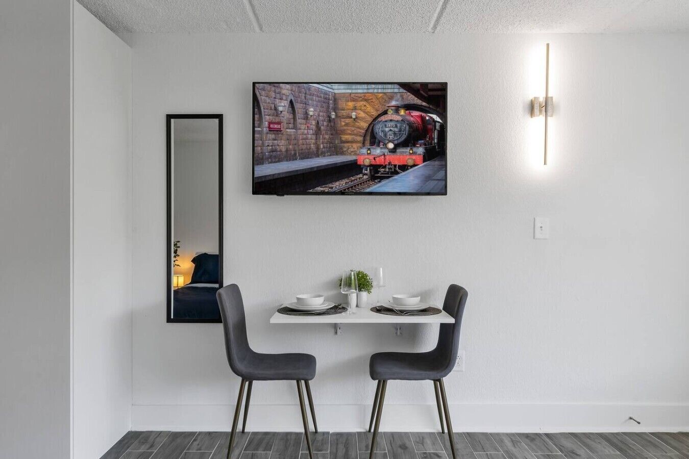 Un comedor moderno con una televisión de pantalla plana en la pared, dos sillas, una mesita y un espejo.