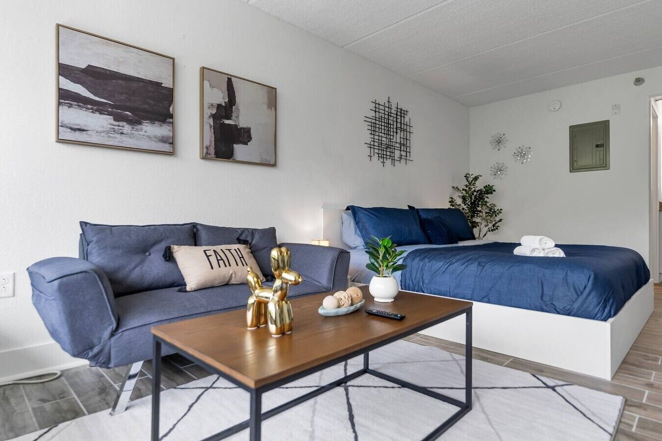 A modern bedroom with a blue sofa, a bed with blue bedding, a wooden coffee table, and a wall with abstract art.