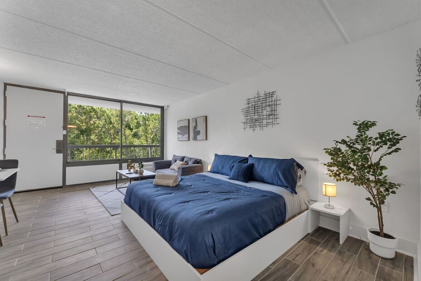 A modern bedroom with a large bed, a desk, and a view of greenery through a sliding door.