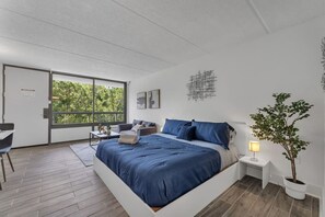 A modern bedroom with a large bed, a desk, and a view of greenery through a sliding door. - Dream Studio 7 Min to Disney + Gym & Parking I Orl (Kissimmee)