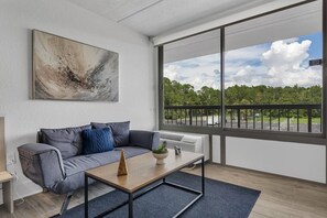 A modern living room with a grey sofa, a coffee table, and a large window offering a view of a parking lot and greenery. - Best Disney Location + Pool & Free Parking | Orl (Kissimmee)