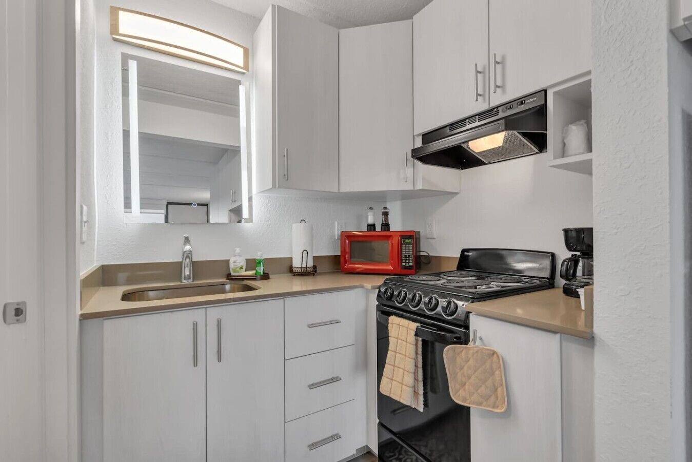 A compact kitchen with white cabinets, a stainless steel sink, a red microwave, a black stove, and a coffee maker.