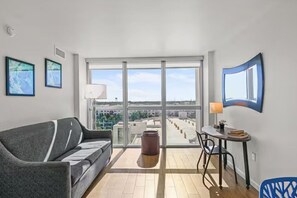 A modern living room with a grey sofa, a small round table, and a chair. There is a large window offering a view of the cityscape. - Cozy Retreat with Stunning Water View | Fort Myers (Fort Myers)