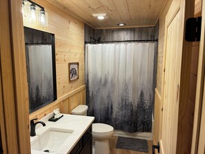 Un baño con paredes de madera, un lavabo blanco, un inodoro y una cortina de ducha con diseño de bosque.