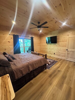 Un dormitorio con paneles de madera, una cama, un ventilador de techo, un televisor montado en la pared y vistas al exterior a través de una ventana.