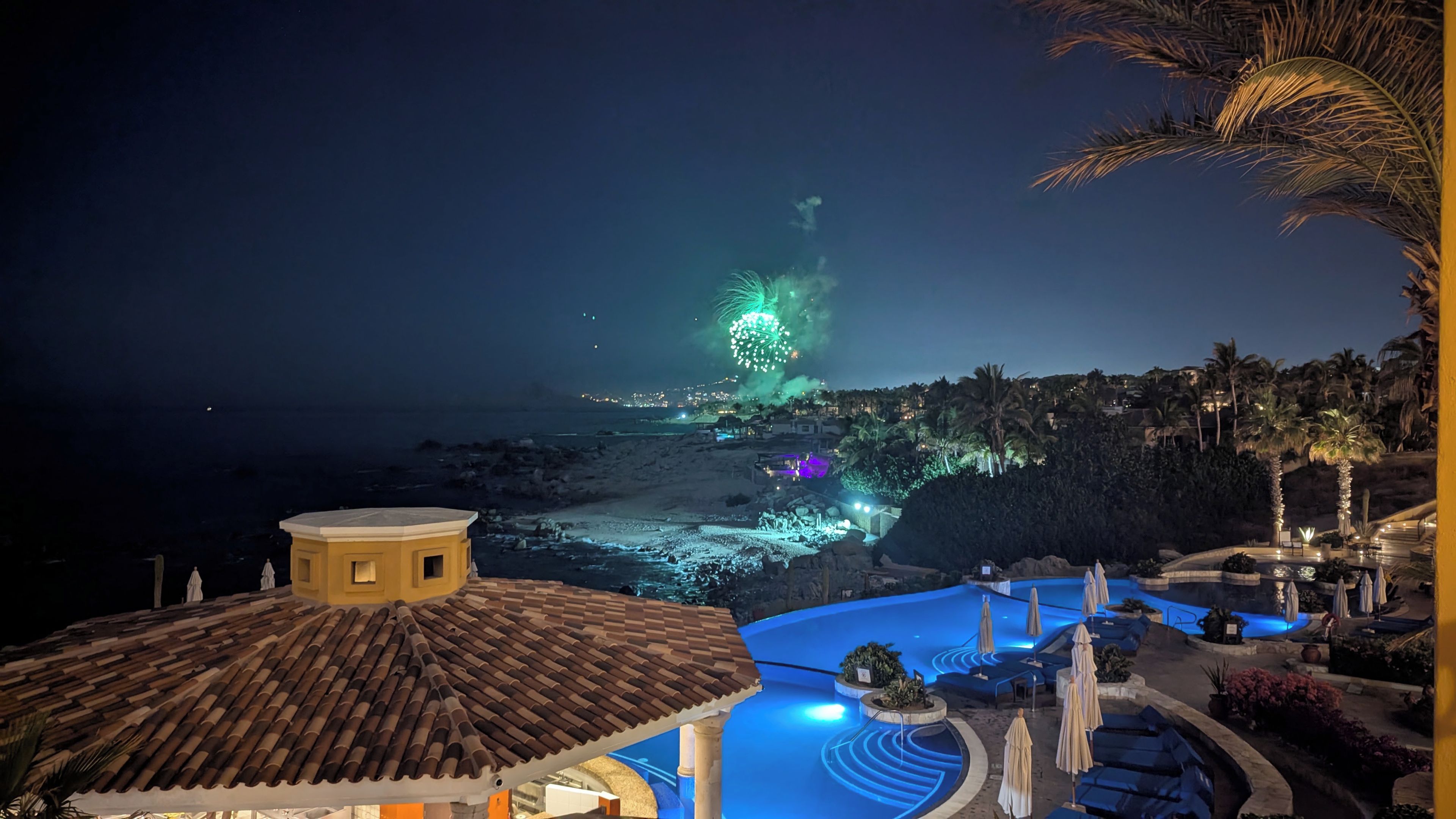 Resort com piscina, palmeiras e vista para o oceano à noite.