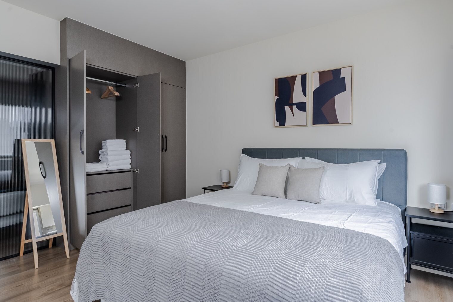 A modern bedroom with a large bed, a wardrobe, a bedside table, and abstract wall art.