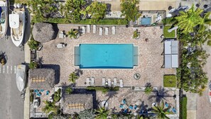 Eine Vogelperspektive zeigt einen Resortkomplex mit einem zentralen Swimmingpool, Liegestühlen und Palmen.