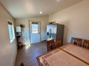 Uma sala de jantar com mesa e cadeiras, geladeira e porta que dá para outro cômodo.
