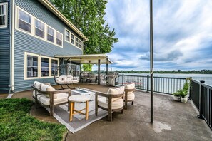 Een terras met tuinmeubilair, een hangstoel en een barbecueplaats met uitzicht op een waterpartij.