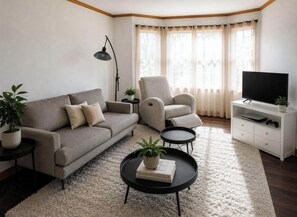 Een woonkamer met een bank, fauteuil, salontafel, tv-meubel en een groot raam met gordijnen.