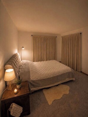 Een slaapkamer met een bed, een nachtkastje met een lamp, een klein plantje en een bont tapijt.